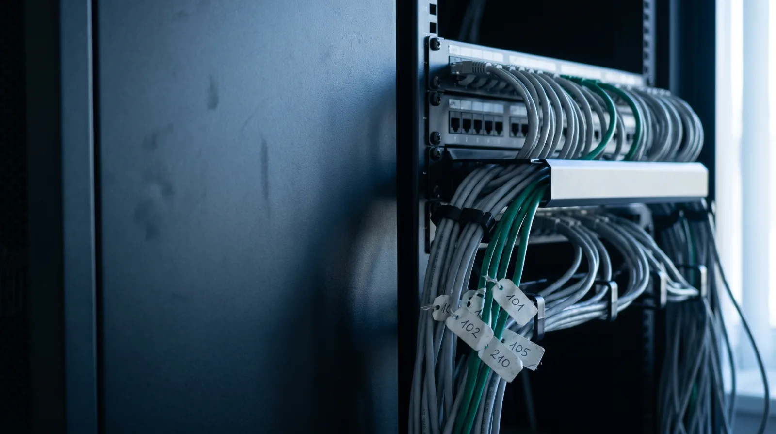 Close-up of a patch panel in a server rack with grey patch cables, a few green accent cables, and hand-written number tags.
